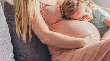 gravid kvinne i sofaen med en liten jente som koser på magen hennes.